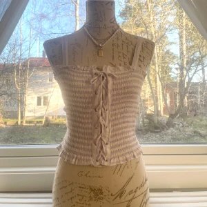 Ljuslila croppad tubtopp med snörning - Säljer en ljuslila och vit randig croppad tubtopp med smock och snörning framtill. Toppen har tunna axelband och volangkant nedtill för en söt look. Perfekt för varma dagar och enkel att styla med jeans eller kjol. är stretchig så passar storlek 32-36.
