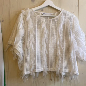 Vit fransig topp från ZARA - Säljer en vit topp från ZARA med fransdetaljer och grovstickad struktur. Modellen är boxig och har korta ärmar. Perfekt för dig som vill sticka ut med en unik och trendig look.
