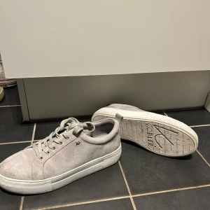Ljusgrå sneakers från K.Cobler - Snygga ljusgrå sneakers från K.Cobler i mocka med vita sulor och snörning. Skorna har en klassisk och stilren design som passar till många outfits. Perfekta för dig som gillar en clean och enkel look.