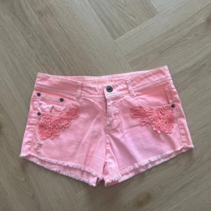 Rosa jeansshorts med spetsdetaljer - Säljer ett par ljusrosa jeansshorts med fransig kant och broderade spetsdetaljer på framfickorna. Shortsen har låg midja och klassisk femficksdesign. Perfekta för varma dagar!