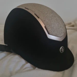 Svart och glittrig hjälm från BR - Snygg och säker hjälm från BR i gammal modell med guldigt glitter. Storlek 52-54. 💕💗 Hjälmen har en justerbar rem och är CE-certifierad för extra säkerhet. Ser ren och ny ut förutom en liten defekt (fläck) på ena sidan. Läder remmarna är ny putsade. 