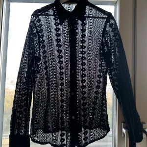 Svart spetsmönstrad skjorta från Our Legacy - Säljer en svart skjorta från Our Legacy med genombrutet spetsmönster och klassisk krage. Skjortan har knappar framtill och långa ärmar.