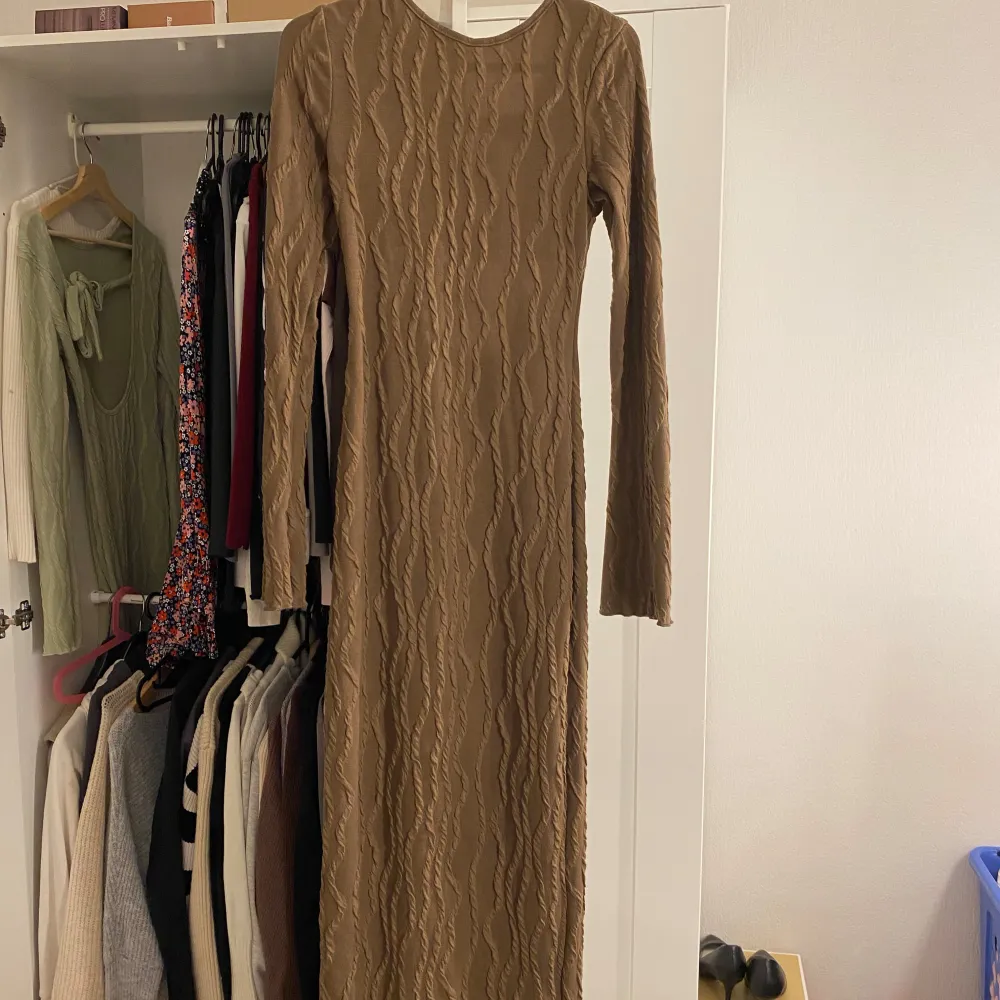 Säljer en brun långklänning med lång ärm och snygg struktur i tyget. Klänningen har ett djupt rundat bröstparti med knyt framtill och är figurnära i modellen. Perfekt för dig som gillar stilrena och trendiga plagg. Storlek Xs (nytt skick). Mekot.