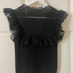 Svart volangtopp från Zara - Säljer en svart topp från Zara med volangdetaljer på axlarna och över bröstet. Toppen är ärmlös och har en lätt transparent mesh upptill. Perfekt för dig som vill ha något trendigt och snyggt!