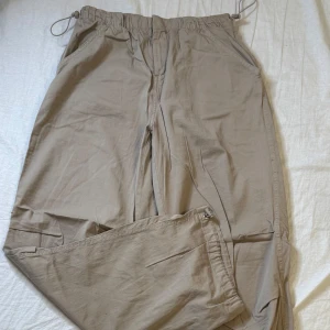 Beige parachute pants från Pull&Bear - Säljer ett par beige cargopants från Pull&Bear med dragsko i midjan och dragkedja. Byxorna har flera fickor och är perfekta för en avslappnad stil. Materialet känns lätt och bekvämt. Pris kan diskuteras