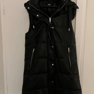 Svart täckväst med huva från Zara i konstläder - Snygg svart väst från Zara med dragkedja och knappar framtill. Västen har en hög krage, huva och fickor med dragkedjor. 🖤