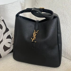 Svart axelväska - Elegant svart axelväska i skinn med justerbar rem och guldiga detaljer. Perfekt för att ge en lyxig touch till din outfit. Den har en stilren design med det ikoniska YSL-emblemet i guld på framsidan. 