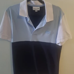 Blå pikétröja från Lacoste - Snygg blå och vit pikétröja från Lacoste med klassisk krage och knappar. 🐊