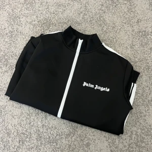 Svart träningsjacka från Palm Angels - Snygg svart träningsjacka från Palm Angels med vita detaljer. Jackan har en dragkedja framtill och vita ränder längs ärmarna. Perfekt för en sportig look. (Ny pris 6400)