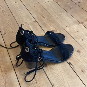 Svarta klackskor med snörning - Snygga svarta klackskor med öppen tå och snörning runt ankeln. Skorna har en stabil klack och är perfekta för en stilren look. De är tillverkade i mocka och har metallringar för snörningen.