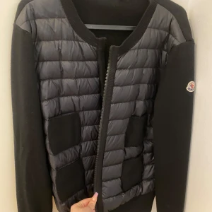 Svart cardigan från Moncler - Säljer en svart cardigan från Moncler med quiltade partier framtill och på axlarna. Jackan har långa ärmar i stickat material och två fickor framtill. Klassisk Moncler-logga på ena ärmen och dragkedja framtill. Kvitto finns. Liten i storleken, är mer som en M. Nypris 9500