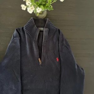Mörkblå stickad tröja med dragkedja från Polo Ralph Lauren - (OBS VET EJ VARFÖR SEN SER SÅ SMUTSIG UT PÅ BILD ÄR EJ DET I VERKLIGHETEN) Säljer en mörkblå stickad tröja från Polo Ralph Lauren med halv dragkedja och hög krage. Tröjan har ett rött broderat logomärke på bröstet och ribbade detaljer vid krage och ärmslut. Perfekt för lager-på-lager-stil.