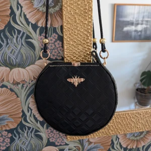 Svart quiltad rund axelväska med gulddetaljer - Säljer en svart rund axelväska med quiltat mönster och snygga gulddetaljer. Väskan har ett dekorativt guldfärgat bi framtill och dragkedja upptill. Axelremmen är svart med guldfärgade metalldetaljer. Perfekt accessoar för att lyfta din outfit.