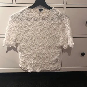 Vit topp från Vero Moda - Säljer en vit blus från Vero Moda med ett unikt texturerat mönster. Blusen har korta ärmar och en lös passform, perfekt för en stilren look. Tillverkad i ett lätt och luftigt material. Använd fåtal gånger, i storlek 36/S