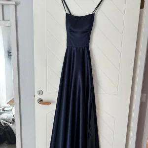 Marinblå balklänning med smala axelband - Marinblå balklänning i satin med smala axelband och snörning i ryggen. Klänningen har en figurnära överdel och ett utsvängt fall, perfekt för bal eller festliga tillfällen. En defekt vid baksida som inte syns så mycket, en tråd har hoppat ur som säkert går att fixa!!💗