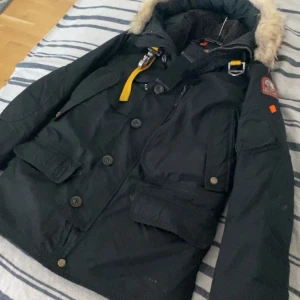  Parajumpers jacka - Säljer en svart parkajacka från Parajumpers med avtagbar huva och pälsdetalj. Jackan har flera fickor, dragkedja och knappar framtill samt en gul rem vid kragen. Perfekt för kalla dagar.