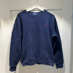 Mörkblå sweatshirt från Polo Ralph Lauren - Säljer en klassisk mörkblå sweatshirt från Polo Ralph Lauren med det ikoniska röda logotypbroderiet på bröstet. Tröjan har en rund halsringning och långa ärmar. Perfekt för en avslappnad stil.