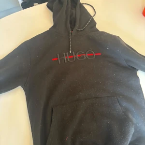 Svart hoodie från Hugo Boss - Säljer en stilren svart hoodie från Hugo Boss med röd och svart logga på bröstet. Hoodien har en klassisk design med dragsko i huvan och en stor magficka framtill. Perfekt för en avslappnad look.