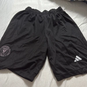 Svarta Inter Miami x Adidas shorts - Svarta sportiga shorts från Adidas med Inter Miami CF-logga på ena benet och Adidas vita logga på det andra. Elastisk midja för skön passform, perfekt för fotboll eller träning. Lätta och luftiga i syntetmaterial. (De är ganska lågmidjade också)