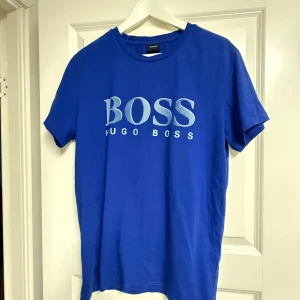 Blå Hugo Boss t-shirt med tryck - Snygg blå t-shirt från Hugo Boss med stort vitt BOSS-tryck på bröstet. Klassisk rund halsringning och korta ärmar. Skön passform och tillverkad i mjuk bomull, perfekt till jeans eller shorts.