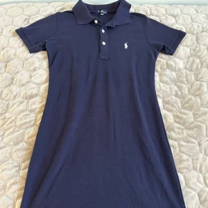 Marinblå pikéklänning från Ralph Lauren - Klassisk marinblå pikéklänning från Ralph Lauren med krage, korta ärmar och knappar framtill. Klänningen har en slim passform och är tillverkad i mjuk bomull. 