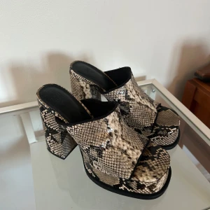 Ormskinnsmönstrade platåklackar H&M - Chunky platåklackar från H&M i ormskinnsmönstrat syntetmaterial. Skorna har öppen tå, bred rem över foten och hög blockklack. Färgerna går i beige, svart och grått för en edgy vibe. Perfekta för dig som vill sticka ut med coola detaljer.