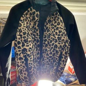 Leopardmönstrad pufferjacka från North - Snygg pufferjacka från North med leopardmönstrad front i beige och svart. Ärmarna är svarta och jackan har dragkedja framtill samt två dragkedjefickor. Perfekt för dig som vill sticka ut med en cool och trendig look.