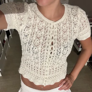 Vit virkad blus med pärlknappar - Supersöt vit virkad blus med korta ärmar och genombrutet spetsmönster. Toppen har pärlknappar framtill och en vågig kant nertill som ger en romantisk vibe. Perfekt för dig som gillar vintage-inspirerade detaljer och vill ha något unikt i garderoben.