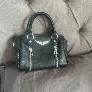 Svart handväska med nitar och dragkedjor - Cool svart handväska i skinn med två rejäla handtag, silverfärgade dragkedjor framtill och små nitar längs kanterna. Väskan har en snygg vinge i silver på framsidan och en robust, lite boxig form. Perfekt för dig som gillar detaljer och rockig stil. Inte äkta