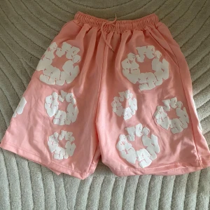 Denim Tears Shorts - Säljer ett par ljusrosa shorts med vita blommiga tryck och elastisk midja med snörning. De är loose fit och perfekta för sommaren. Materialet känns mjukt och svalt. Riktigt sköna och snygga för chill dagar. Pris kan diskuteras🙌🙌