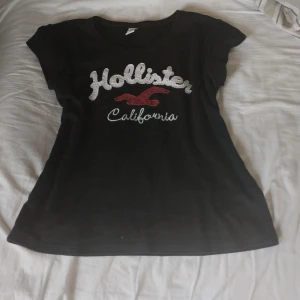 Svart Hollister t-shirt med brodyr - Svart t-shirt från Hollister med vit broderad logga och röd fågel på bröstet. Modellen är figurnära med rund hals och korta ärmar. Materialet känns mjukt och skönt, perfekt till jeans eller shorts. Klassisk och stilren med tydlig California-vibe. Den är i storlek m men passar mig som bär xs💗Skriv för funderingar❤️