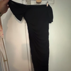 Svart offshoulder midiklänning - Snygg svart långklänning med offshoulder-design och draperad effekt längs hela klänningen. Tunnare material som ger ett elegant fall och markerar siluetten. Perfekt för dig som vill sticka ut med en stilren och trendig look.