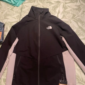 Svart Phlego track jacket från The North Face - Track jacket från The North Face i svart med gråa paneler längs sidorna och ärmarna. Denna modell tillverkas inte längre och är slutsåld överallt. Helt oanvänd med alla lappar kvar. Jackan har hög krage, hel dragkedja framtill och två fickor med dragkedja. Logga på bröstet och ärmen. Perfekt för en sportig och clean look.