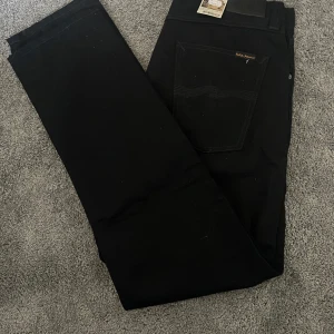 Svarta jeans från Nudie Jeans Co - Snygga svarta jeans från Nudie Jeans Co med klassisk femficksdesign och raka ben. Jeansen har svarta sömmar, silverfärgade nitar och knapp med logga. Jeansen är helt oanvända och har prislappen kvar. Hör av er vid frågor