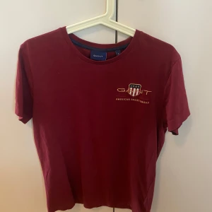 Vinröd t-shirt från GANT, S - Vinröd t-shirt från GANT med broderad logga och sköld på bröstet. Klassisk rund halsringning och korta ärmar. Tillverkad i mjuk bomull som känns skön mot huden. Perfekt för dig som gillar stilren och sportig vibe. Ny pris mellan 500-900