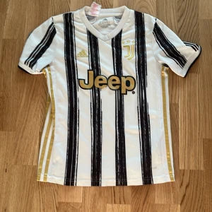 Juventus Adidas fotbollströja 11-12 år - Juventus matchtröja från Adidas i vitt med svarta och guldiga detaljer. Tröjan har svarta ränder, guldiga logotyper och Jeep-tryck på bröstet. Tillverkad i lätt och ventilerande AEROREADY-material, perfekt för fotbollsträning eller att bära på läktaren.