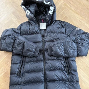 Svart Moncler dunjacka med huva - Snygg svart dunjacka från Moncler med huva och dragkedja framtill. Jackan är quiltad och har Moncler-logga på ärmen samt coolt serietryck på insidan. Perfekt för kalla dagar och har en riktigt clean och puffig look. STORLEK 5