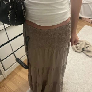 Beige långkjol med smock i midjan - Superskön beige långkjol med smockad midja och volangiga lager. Kjolen har en loose fit och är perfekt för dig som gillar en avslappnad och trendig stil. Passar lika bra med crop top som med stickad tröja. Enkel att matcha och riktigt flowy.