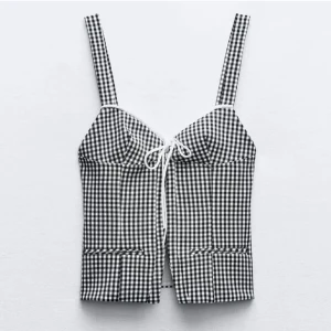 Rutig korsettopp med snörning - Supersnygg korsettopp med svartvitt rutigt mönster och axelband. Toppen har en söt snörningsdetalj framtill och markerade sömmar som ger en cool siluett. Perfekt för dig som vill sticka ut med en trendig och lekfull look. Säljer då den inte passade mig