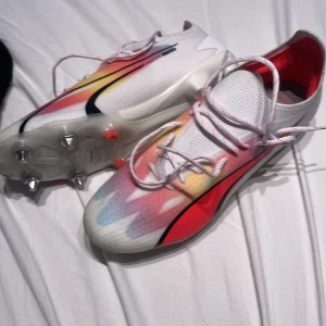 Puma fotbollsskor i färgglad design - Snygga fotbollsskor från Puma  med vit bas och färgskiftningar i rött, gult och blått. Skorna har snörning, svart swoosh och metall-dobbar för bästa grepp på planen. Ovandelen är i syntetmaterial och designen är riktigt modern och sportig. skruvdobbar, aldrig använda