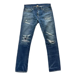 Blå slitna jeans med raka ben - Säljer ett par blå jeans med coola slitningar och hål på benen. Jeansen har raka ben och klassisk femficksdesign. Perfekta för en avslappnad streetstil och passar till sneakers eller boots.