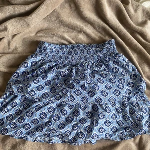 Blå mönstrad volangkjol med resårmidja - Supersöt volangkjol i blått med vitt och mörkblått mönster. Kjolen har bred resår i midjan för skön passform och är gjord i ett lätt och luftigt material, perfekt för varma dagar. Snygg att matcha med en enkel topp eller hoodie. Aldrig andvänt. Pris kan diskuteras.