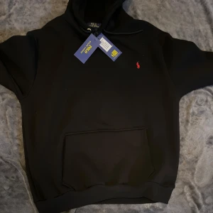 Svart hoodie från Polo Ralph Lauren - Svart hoodie från Polo Ralph Lauren med klassisk röd broderad logga på bröstet. Tröjan har huva med snörning och en stor magficka framtill. Tillverkad i mjuk bomull, perfekt för en chill och stilren look. Vill sälja snabbt pris kan diskuteras