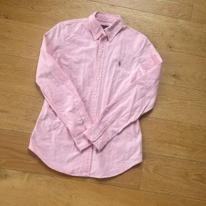 Ralph Lauren Skjorta (NY SKICK) - Säljer en klassisk rosa skjorta från Ralph Lauren i SLIM FIT med button-down krage och broderad logga på bröstet. Skjortan har lång ärm, normal passform och är tillverkad i mjuk bomull. Perfekt för dig som gillar stilrena och tidlösa plagg.