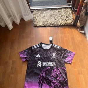Liverpool x Adidas svart/lila fotbollströja - Snygg Liverpool fotbollströja från Adidas i slim fit. Tröjan är svart med lila och rosa toner, har korta ärmar och ett unikt grafiskt mönster med en stor fågel på framsidan. Klassiska Adidas-ränder på axlarna och Liverpool FC-logga på bröstet. Materialet är lätt och andas.