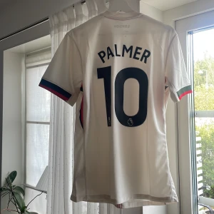Chelsea Palmer 10 bortatröja Nike M - Chelsea FC bortatröja med Palmer 10-tryck. Vit med tunna ränder i rött och grönt, klubbmärke på bröstet och Nike-logga. Ärmarna har blå, röda och gröna detaljer. Tillverkad i Dri-FIT-material för bästa komfort. Snygg och modern passform.