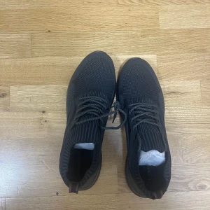 Svarta sneakers från Pier One - Svarta sneakers från Pier One med stickad ovandel och snygga detaljer. Skorna har snörning, ribbade partier runt öppningen och en platt, mönstrad sula för bra grepp. Perfekta för en clean och sportig look.