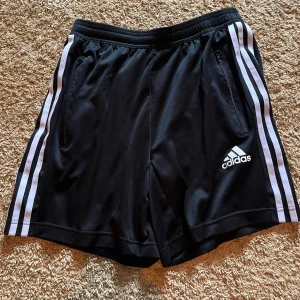 Svarta Adidas shorts med vita ränder - Klassiska svarta Adidas shorts med vita ränder längs sidorna och logga framtill. Elastisk midja och dragkedjeförsedda fickor. Perfekta för träning eller chill, obs funkar även som badbyxor. Tillverkade i lätt polyester för maximal komfort. Nypris 319kr! 