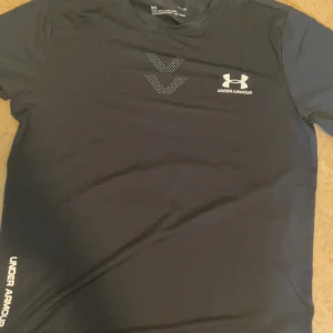 Svart tränings-tshirt från Under Armour - Svart kortärmad t-shirt från Under Armour med vit logga på bröstet och diskreta prickdetaljer. Gjord i ett lätt och snabbtorkande syntetmaterial, perfekt för träning. Snygg, sportig look med rund hals och tryck även på ärmen.