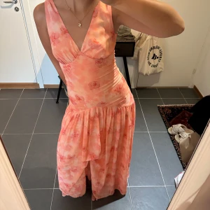 Adoore klänning - Superfin långklänning i ljusrosa och persikofärgade toner med blommigt mönster. Djup v-ringning fram och öppen rygg, ärmlös modell och mjukt, följsamt material. Perfekt för sommarens alla tillfällen!